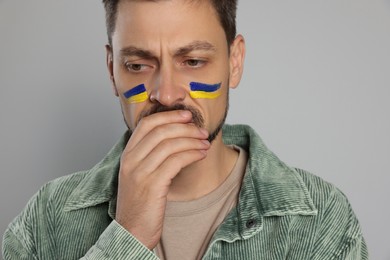 Photo of Sad man with drawings of Ukrainian flag on face against light grey background, closeup