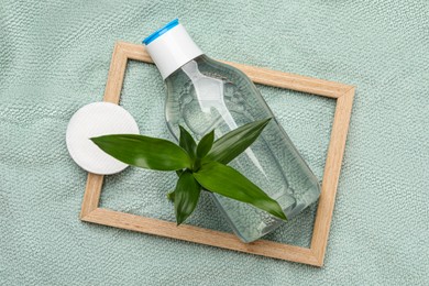 Bottle of micellar cleansing water, cotton pads and green plant on turquoise towel, flat lay