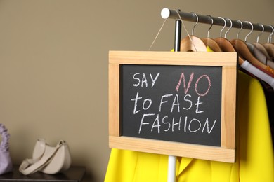 Photo of Small chalkboard with phrase SAY NO TO FAST FASHION and different stylish clothes on rack indoors, space for text