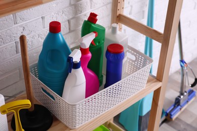 Plunger and basket with detergents on wooden shelf near white brick wall indoors