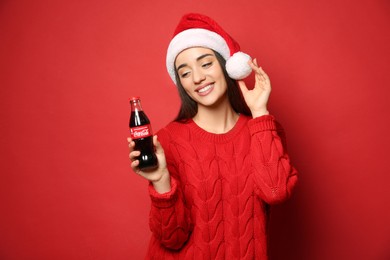 MYKOLAIV, UKRAINE - JANUARY 27, 2021: Young woman in Christmas hat holding bottle of Coca-Cola on red background