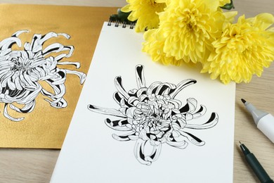 Drawings of chrysanthemum with flowers and pens on wooden table, closeup