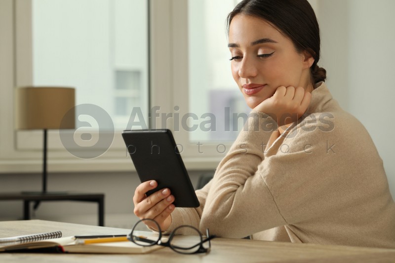 Young woman using e-book reader at wooden table indoors