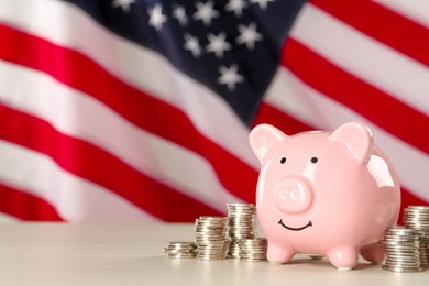 Piggy bank and coins on white wooden table against American flag, space for text