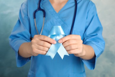Photo of Doctor holding light blue awareness ribbon on color background, closeup