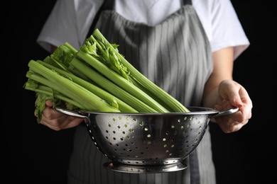 Photo of Woman holding colander with fresh green celery on black background, closeup