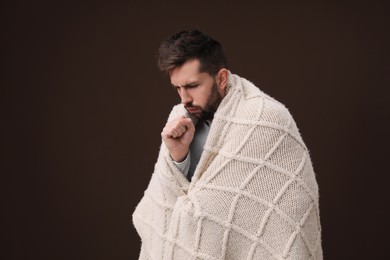 Man wrapped in blanket coughing on brown background. Cold symptoms