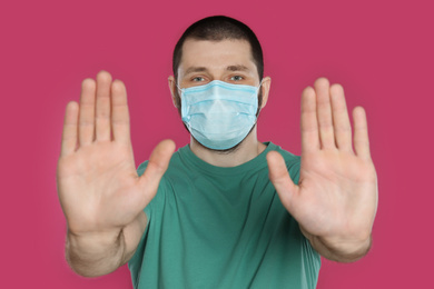 Man in protective mask showing stop gesture on pink background. Prevent spreading of coronavirus