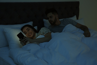 Distrustful young man peering into girlfriend's smartphone in bed at night