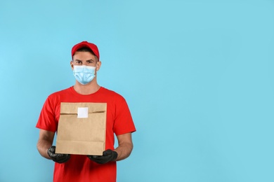 Courier in medical mask holding paper bag with takeaway food on light blue background, space for text. Delivery service during quarantine due to Covid-19 outbreak