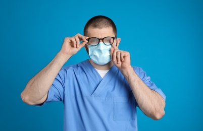 Doctor wiping foggy glasses caused by wearing disposable mask on blue background. Protective measure during coronavirus pandemic