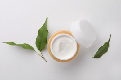 Photo of Jar of face cream and green leaves on white background, flat lay