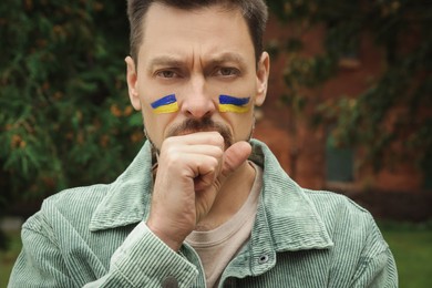 Photo of Sad man with drawings of Ukrainian flag on face outdoors, closeup