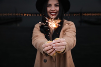 Woman in hat holding burning sparkler near building, focus on hands