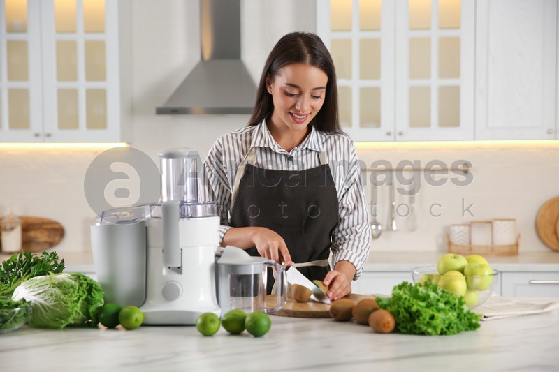 Photo of Young woman cutting fresh kiwi for juice at table in kitchen