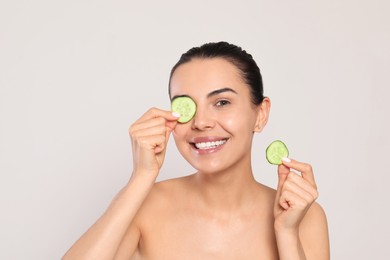 Beautiful young woman putting slices of cucumber on eyes against light grey background