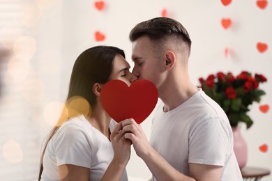 Photo of Lovely couple kissing behind red paper heart indoors. Valentine's day celebration