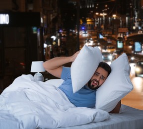 Man covering ears with pillows in bed and beautiful view of night cityscape on background. Poor sleep because of urban noise