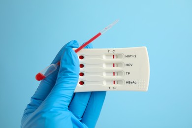 Doctor holding disposable express test for hepatitis and pipette with blood on light blue background, closeup