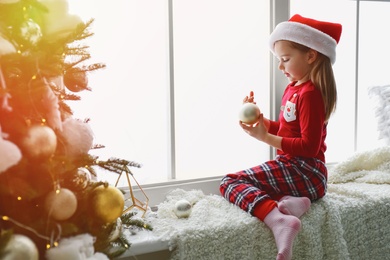 Photo of Cute little girl in Santa hat holding Christmas ball on window sill at home