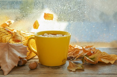 Cup of hot drink with marshmallows and autumn leaves near window on rainy day. Cozy atmosphere