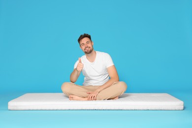 Smiling man sitting on soft mattress and showing thumb up against light blue background