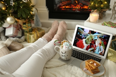 MYKOLAIV, UKRAINE - DECEMBER 23, 2020: Woman watching The Grinch movie on laptop near fireplace at home, closeup. Cozy winter holidays atmosphere