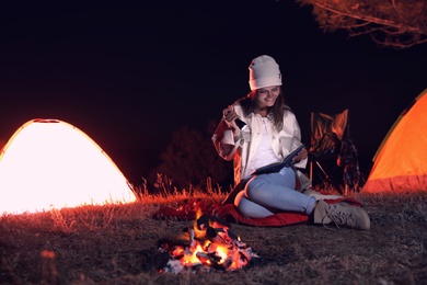 Young woman with flashlight reading book near bonfire at night. Camping season