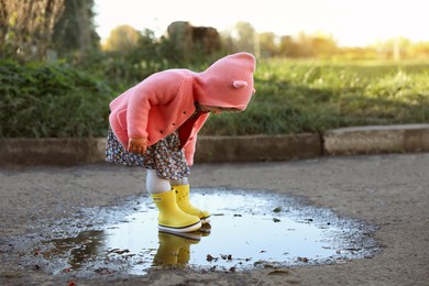 Little girl wearing rubber boots standing in puddle outdoors, space for text. Autumn walk