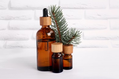 Bottles of essential oil and pine branch on white wooden table near brick wall