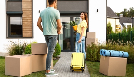 Photo of Couple walking to their new house with moving boxes and household stuff