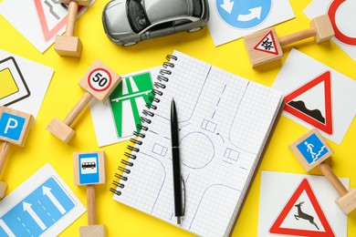 Many different road signs, notebook with sketch of roundabout and toy car on yellow background, flat lay. Driving school