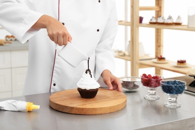 Female pastry chef pouring chocolate sauce onto cupcake at table in kitchen, closeup