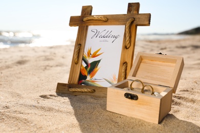 Beautiful wedding invitation and wooden box with gold rings on sandy beach