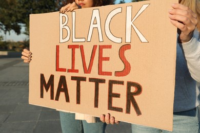 Women holding sign with phrase Black Lives Matter outdoors, closeup. Racism concept