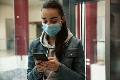 Young woman with disposable mask and smartphone in elevator. Dangerous virus