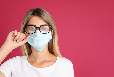 Woman wiping foggy glasses caused by wearing disposable mask on pink background, space for text. Protective measure during coronavirus pandemic