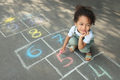Photo of Little African American girl drawing hopscotch with chalk on asphalt outdoors. Happy childhood