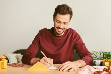 Photo of Happy man writing message in greeting card at wooden table in room