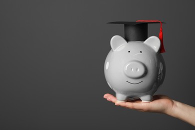 Photo of Woman holding piggy bank and graduation cap against dark grey background, closeup with space for text. Scholarship concept
