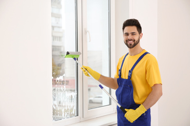 Professional young janitor in uniform cleaning window indoors