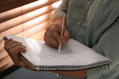 Man drawing portrait with pencil in notepad indoors, closeup