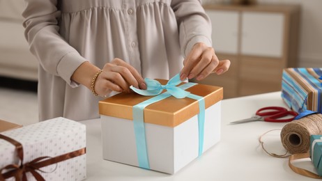 Woman decorating gift box with ribbon at white table indoors, closeup
