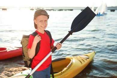 Happy boy with paddle near kayak on river shore. Summer camp activity
