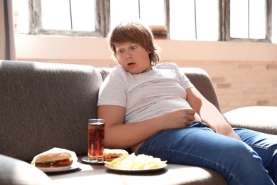 Emotional overweight boy with fast food on sofa at home