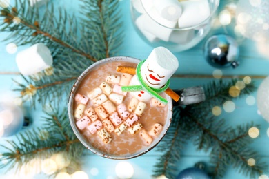 Funny marshmallow snowman in cup of hot drink on light blue wooden table, top view. Bokeh effect 