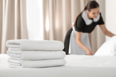 Young maid making bed in hotel room, focus on stack of towels