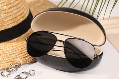 Stylish sunglasses in black leather case near straw hat and earrings on white table