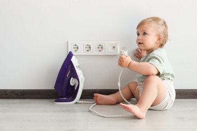 Photo of Cute baby playing with electrical socket and iron plug at home. Dangerous situation
