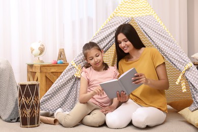 Mother and daughter reading book near toy wigwam at home